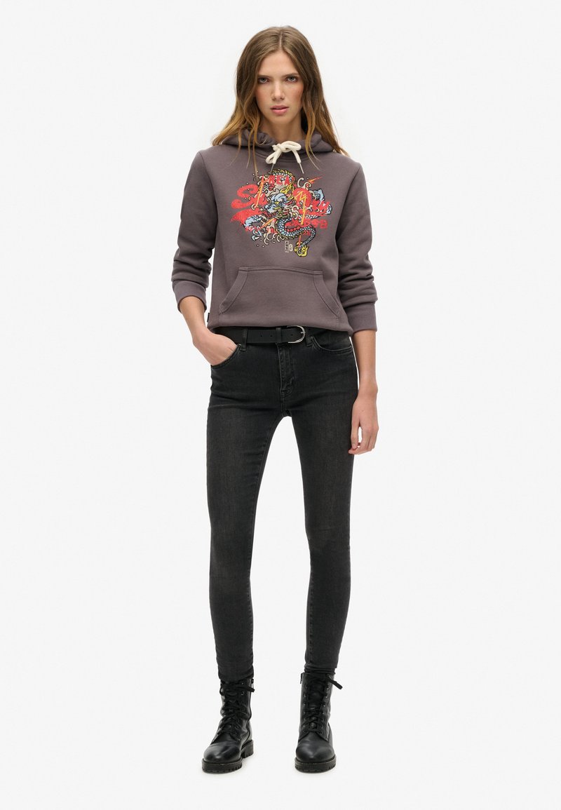 Kobieta w szarej bluzie z kapturem z graficznym wzorem smoka, w czarnych obcisłych dżinsach, z czarnym paskiem i czarnymi sznurowanymi butami, stojąca na białym tle.