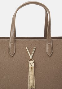 Brūna tekstūrēta rokassoma ar divām rokturiem, zelta aksesuāru ar "V" logotipu un pušķi.