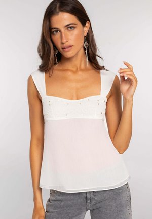 Mujer con camiseta blanca sin mangas con tirantes anchos y bordado decorativo, combinada con vaqueros grises con tachuelas, posando sobre un fondo liso.