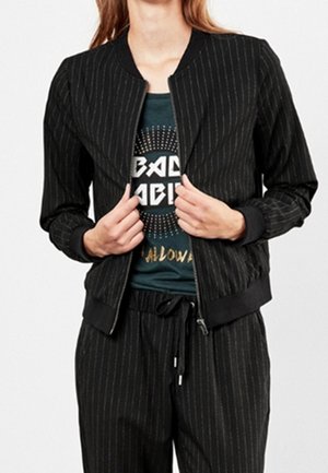 Femme portant une veste zippée noire à fines rayures sur un T-shirt graphique sombre et un pantalon à fines rayures assorti avec taille à cordon.