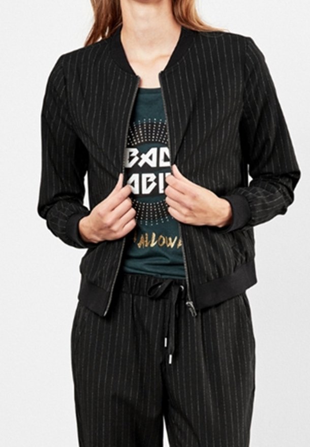 Femme portant une veste zippée noire à fines rayures sur un T-shirt graphique sombre et un pantalon à fines rayures assorti avec taille à cordon.