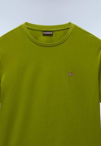 Green cotton t-shirt with a round neckline and short sleeves. Features a small embroidered flag detail on the chest. Soft texture.