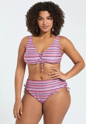 Kvinde med krøllede hår iført en lyserød, mønstret todelt badedragt, der står med den ene hånd på hoften foran en ensfarvet baggrund.