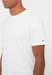 Hombre vestido con una camiseta de manga corta blanca lisa, con un pequeño logo bordado en la manga izquierda y texto sutil en el pecho.