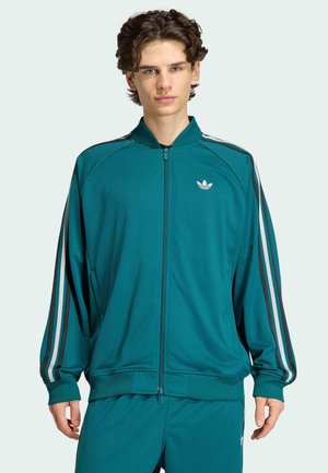 Giovane uomo che indossa una tuta Adidas verde acqua con strisce bianche e nere sulle maniche e sui pantaloni, in piedi davanti a uno sfondo chiaro.