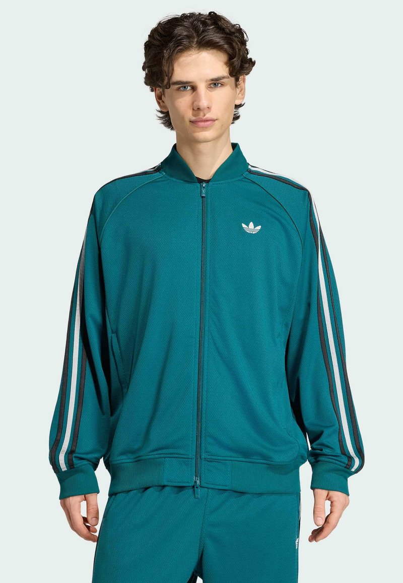 Jeune homme portant un survêtement Adidas bleu sarcelle avec des bandes blanches et noires sur les manches et le pantalon, debout devant un fond clair.
