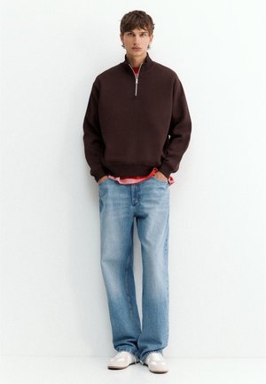 Jeune homme debout, portant un pull marron foncé à fermeture éclair quart, un jean bleu clair, des baskets blanches et une chemise rouge dépassant à l'ourlet.