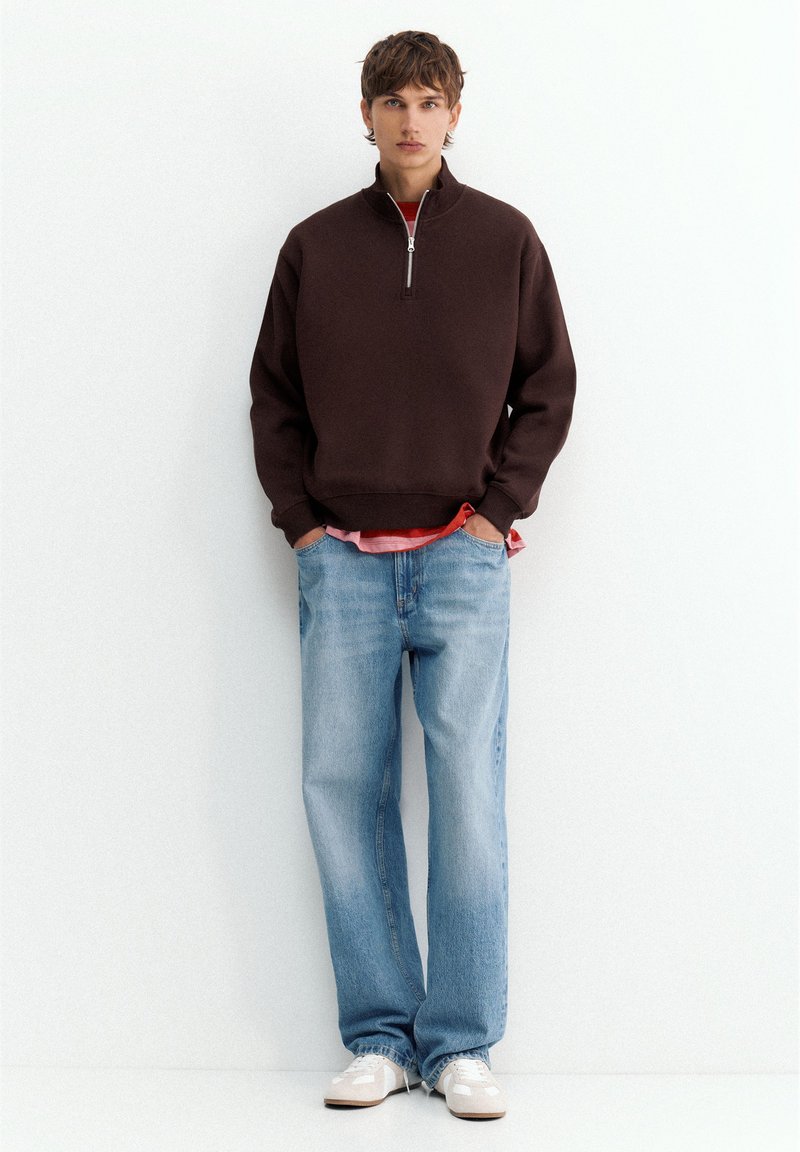 Jeune homme debout, portant un pull marron foncé à fermeture éclair quart, un jean bleu clair, des baskets blanches et une chemise rouge dépassant à l'ourlet.