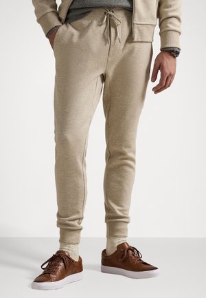 Hombre con joggers con cordón beige, calcetines beige, zapatillas de cuero marrón y chaqueta beige con las manos en los bolsillos, posando sobre un fondo liso.