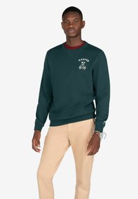 Sudadera de color verde oscuro con un gráfico frontal que presenta el texto "HARPER" y un diseño de oso. Puños y dobladillo acanalados, combinada con pantalones cargo beige.
