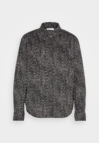 Chemise noire à manches longues avec boutons, motif de points blancs irréguliers et col classique, présentée sur un fond clair uni.