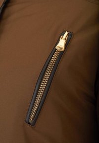 Braune Jacke mit einem auffälligen seitlichen Reißverschluss und goldenen Beschlägen sowie dunklen Einfassungen, aus glattem, strapazierfähigem Stoff gefertigt.