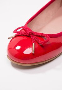 Röda lackläderballettskor med rund tå, med en texturerad röd rosett och gyllene detaljer i ändarna av rosetten.