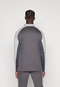 Γκρι αθλητικό μπλουζάκι μακρυμάνικο με ραγκλάν μανίκια. Διαθέτει ανοιχτό γκρι λεπτομέρειες και ψηλό γιακά, κατασκευασμένο από απαλό, ανθεκτικό ύφασμα.