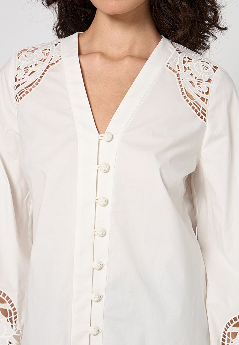 Frau in einer weißen Bluse mit floralem Spitzen-Ausschnitt an Schultern und Ärmeln, V-Ausschnitt und stoffbezogenen Knöpfen vorne.