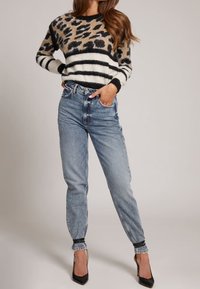 Pull à motif léopard avec des rayures noires et crème, associé à un jean bleu clair taille haute. Le jean a des poignets effilés et des talons noirs.