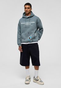 Grijze hoodie met witte tekst en verfspattenaccenten, kangoeroe-pocket, gedragen over een wit T-shirt, gecombineerd met zwarte shorts en sneakers.