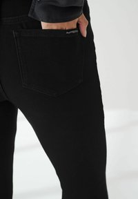 Jeans negros de denim, con un bolsillo trasero que presenta un sutil detalle de logo, textura suave y forma ajustada, mostrados de lado.