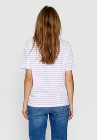 Žena s dlouhými hnědými vlasy, oblečená v bílém tričku s růžovými vodorovnými pruhy a modrými džíny, stojí zády k fotoaparátu na jednolitém pozadí.