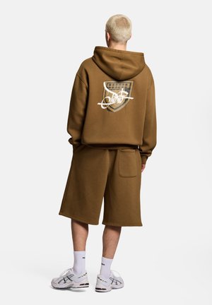Osoba z krótkimi blond włosami, ubrana w brązową bluzę z kapturem z graficzną tarczą i napisem, dopasowane długie szorty, białe skarpetki i białe trampki, widziana od tyłu.