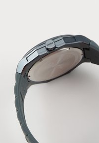 Montre Hugo avec un bracelet en silicone gris, un boîtier métallique, une couronne texturée et un dos argenté réfléchissant. Présente un design minimaliste.