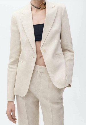 Frau trägt einen beige maßgeschneiderten Anzug mit einreihigem Blazer, passenden Hosen, schwarzem Bandeau-Top und einem Choker mit goldenem Herzanhänger.