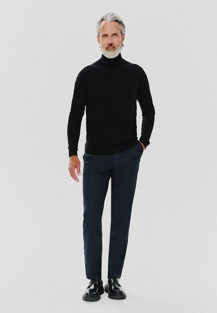 Czarny sweter z golfem, granatowe spodnie, błyszczące czarne buty. Mężczyzna stoi prosto z rękami w kieszeniach, ma szare wąsy i włosy.