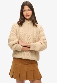 Beige kabelstrik sweater med høj krave og lynlåslukning, parret med en brun plisseret mini nederdel. Struktureret strikkemønster hele vejen igennem.