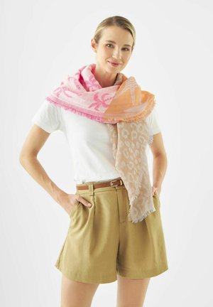 Lächelnde Frau in weißem T-Shirt, khakifarbenen Shorts und einem rosa-orange gemusterten Schal über den Schultern vor weißem Hintergrund.