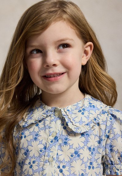 Νεαρή κοπέλα με ανοιχτό καστανά μαλλιά που φοράει μπλε και λευκό φόρεμα με λουλούδια, κοιτάζει προς τα πάνω και χαμογελά ελαφρώς με ουδέτερο φόντο.