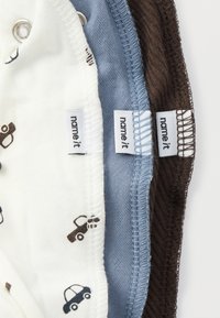 Tres peleles para bebé: uno blanco con estampados de coches, uno azul claro y uno marrón. Cuentan con cierres a presión y tejido de algodón suave. Etiquetas visibles.