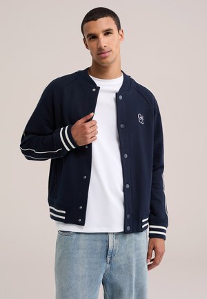 Jeune homme portant une veste varsity bleu marine avec des poignets et un ourlet rayés sur un t-shirt blanc et un jean bleu clair, debout devant un fond uni.
