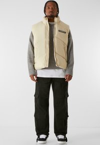 Chaleco acolchado beige con cuello alto, usado sobre un suéter gris, combinado con pantalones cargo negros con múltiples bolsillos y zapatillas oscuras.