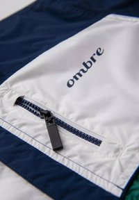 Marinblå och vit vindjacka med dragkedjeficka och "ombre"-logotyp. Har en texturerad tyg och kontrasterande sömdetaljer.
