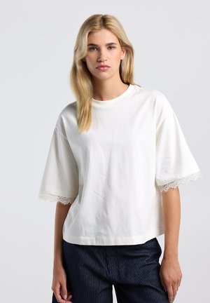 Blonde vrouw die een loszittend wit T-shirt met kanten afwerking aan de mouwen en donkere krijtstreeppantalon draagt, staand tegen een effen grijze achtergrond.