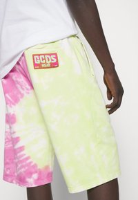Personne portant un short tie-dye rose et jaune pâle avec un patch logo GCDS Wear sur la poche arrière, assorti à une chemise blanche.