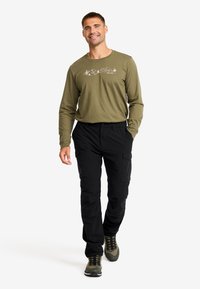 Olive long-sleeve shirt with mountain graphic, paired with black cargo pants. Man standing in a relaxed pose, wearing gray and green shoes.
