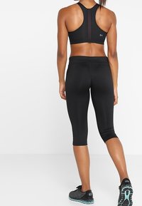 Czarny stanik sportowy z siateczkowym panelem, w połączeniu z czarnymi legginsami ¾. Charakteryzuje się gładkim materiałem i dopasowanym krojem, uzupełniony teksturowanymi butami.