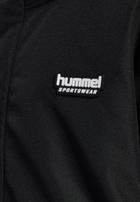 Svart sportjacka med texturerad tyg och en gummibelagd logotyp i patchformat som läser "hummel SPORTSWEAR" på vänster bröst.