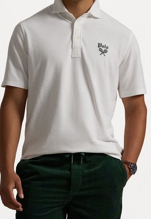 Man draagt een wit poloshirt met het logo "Polo" en gekruiste tennisrackets, gecombineerd met donkergroene corduroy broek en een polshorloge.