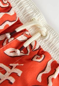 Czerwona tkanina z abstrakcyjnymi kremowymi wzorami, wyposażona w elastyczny pas i biały sznurek ze metalowymi oczkami do regulacji.