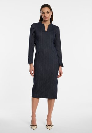 Woman wearing a dark pinstripe knee-length dress with long sleeves and a notch neckline, standing against a plain white background.