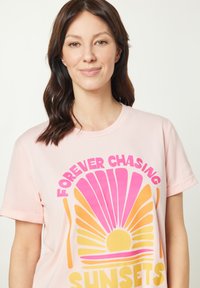 Ljusrosa bomulls-t-shirt med en färgglad grafisk design med sol- och snäcksytor, text som säger "ALLTID EFTER SOLNEDGÅNGAR". Korta ärmar.
