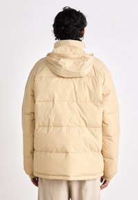 Chaqueta acolchada beige con capucha, diseño de rombos y puños elásticos. El material parece suave, con una textura lisa y un ligero brillo.