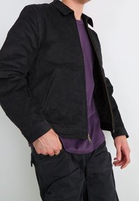 Mężczyzna ubrany w czarną kurtkę z sztruksu na purpurową koszulkę z czarnymi spodniami cargo, z ręką w kieszeni, na jednolitym jasnym tle.