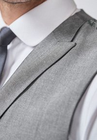Mann in einer grauen Anzüge-Weste, weißem Hemd und gemustertem Krawatte, die den Kragen und den oberen Brustbereich zeigt.