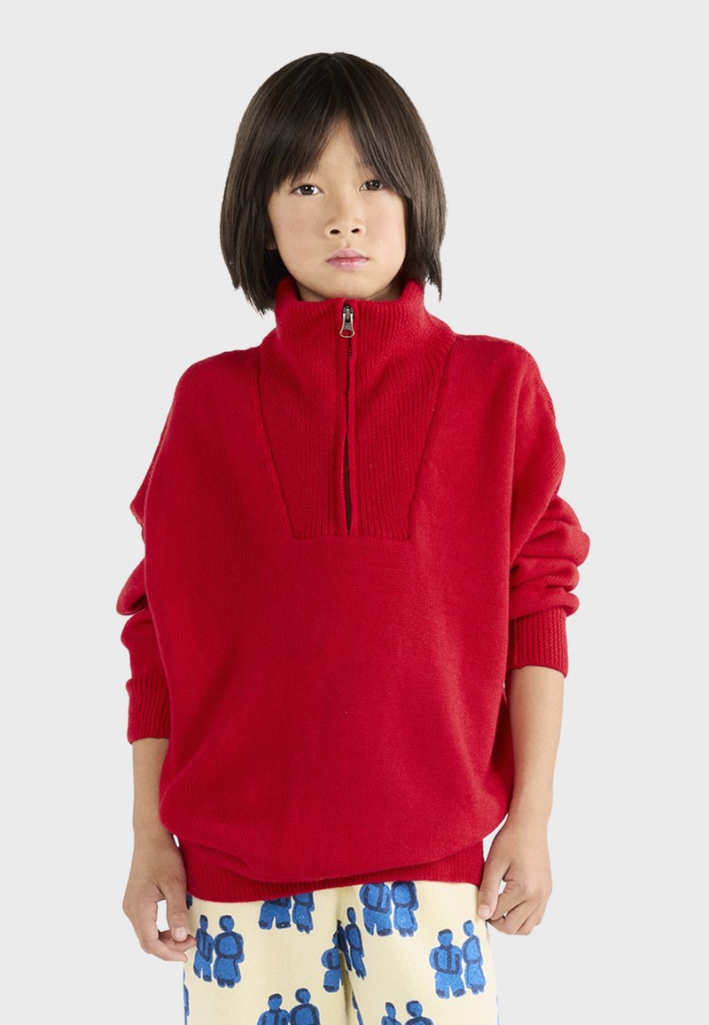 Red knit sweater with high collar and half-zip closure, featuring wide sleeves and a relaxed fit. Worn with patterned light-colored pants.
