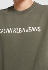 Felpa verde oliva con la scritta bianca "CALVIN KLEIN JEANS". Il testo è in grassetto e si trova sulla parte anteriore del petto. Collo a coste e texture morbida.
