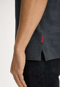 Dunkelgraues Kurzarmhemd aus Baumwolle, mit seitlichen Schlitzen und einem roten Levi's Label für die Markenkennung. Getragen mit schwarzen Jeans.