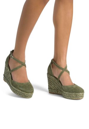 Talons compensés en daim vert avec des brides croisées à la cheville et des détails en corde texturée sur une large semelle compensée. Zone des orteils lisse, design arrondi.